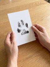 Dos manos sosteniendo una cartulina con la huella real de un perro, ya tomada en tinta negra